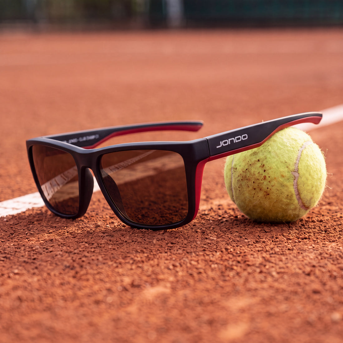 JONDO sunglasses beside a tennis ball on a clay court